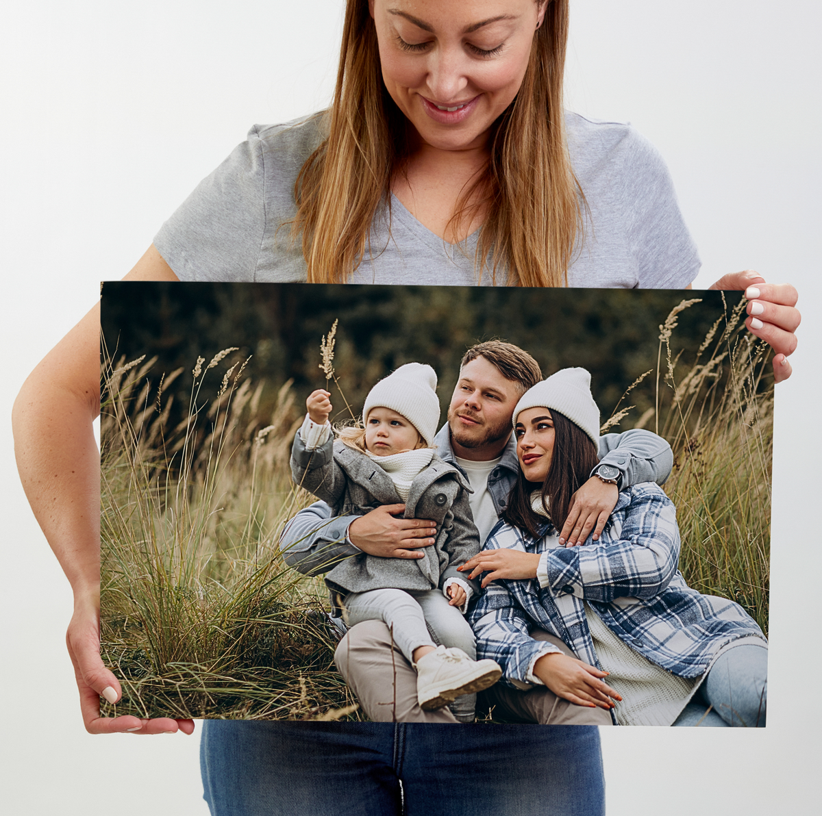A woman holds up a large square custom photo print showing a family of three in a grassy outdoor setting. The woman holding the print is wearing a gray t-shirt and blue jeans, photographed against a clean white background.
